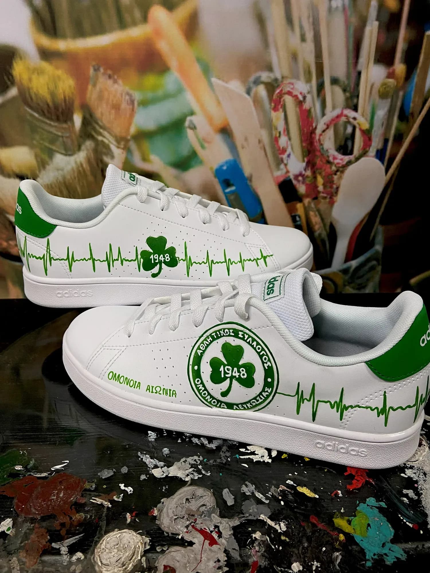 White adidas sneakers hand-painted with the Omonia clover badge and a green heartbeat line