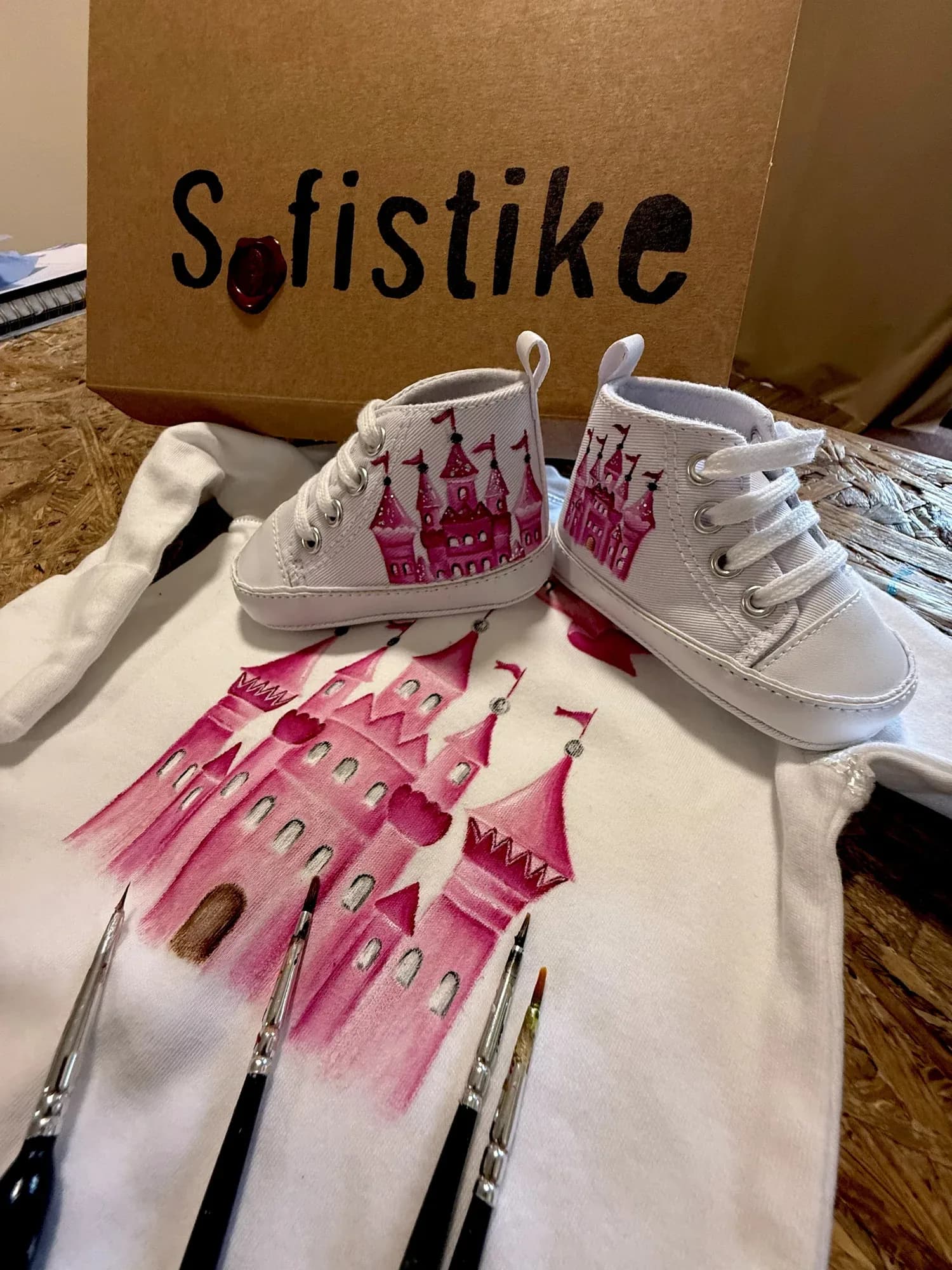 A castle being painted on a baby onesie with brushes, next to baby shoes